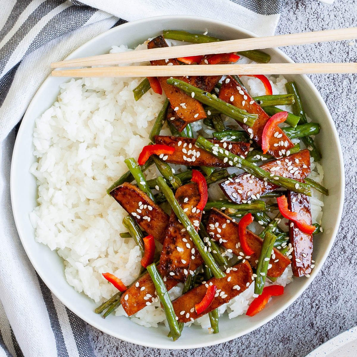 White bowl with white rice topped with thin dark brown tofu slices with charred edges, green beans, chopped red bell pepper and sprinkled with white sesame seeds.