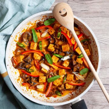 White frying pan with a darkbrown red sauce tofu cubes, sliced bell peppers, sliced green chilies, mint leaves, sliced spring onion, mushrooms slices.