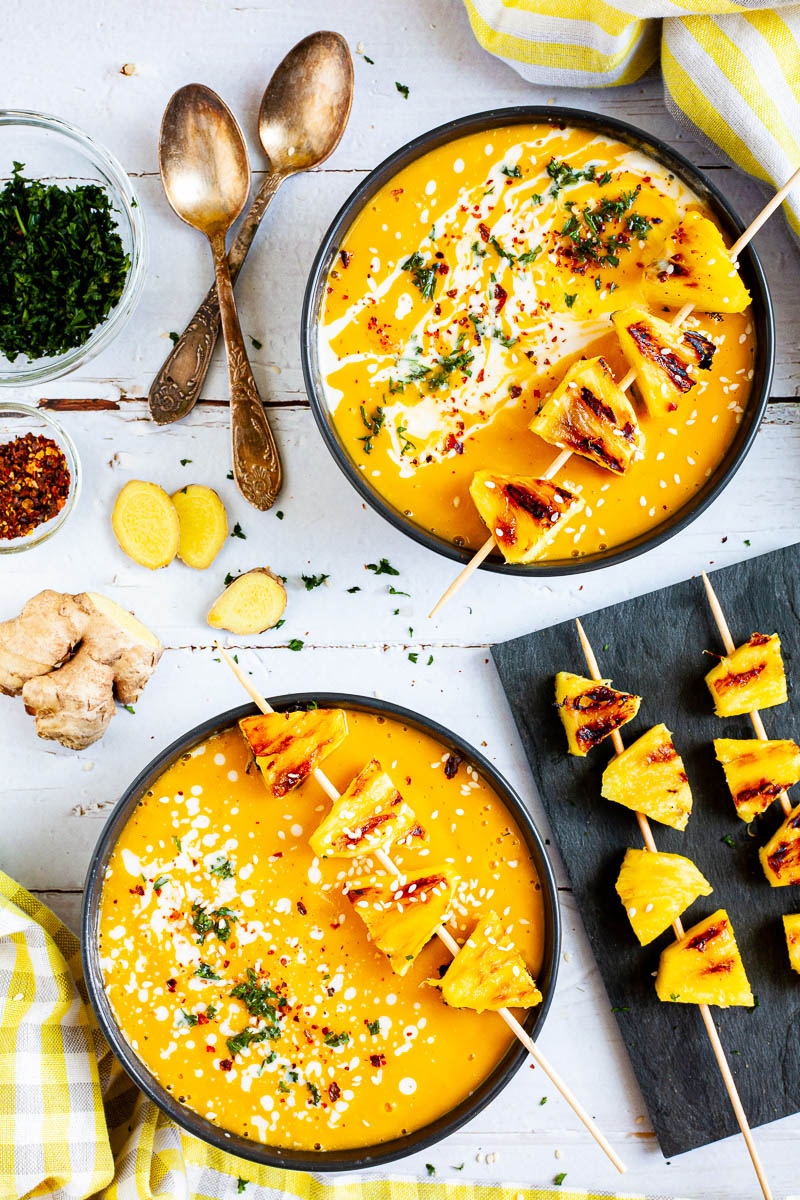 2 black bowls of Yellow cream soup from above topped with charred pineapple wedges, drizzled with a white sauce and sprinkled with chopped green herbs, red flakes and sesame seeds. 2 spoons and the toppings in various bowls are around them.