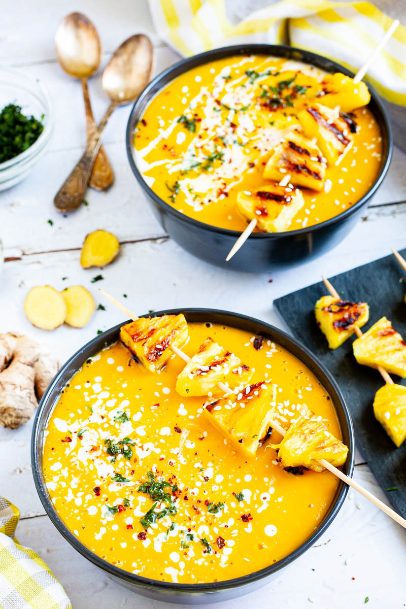 Yellow cream soup in 2 black bowls topped with charred pineapple wedges, drizzled with a white sauce and sprinkled with chopped green herbs, red flakes and sesame seeds.