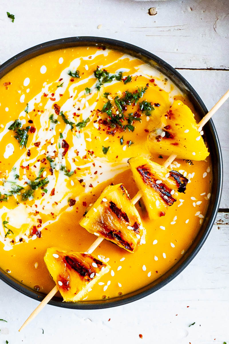 Yellow cream soup topped in a black bowl with charred pineapple wedges, drizzled with a white sauce and sprinkled with chopped green herbs, red flakes and sesame seeds.