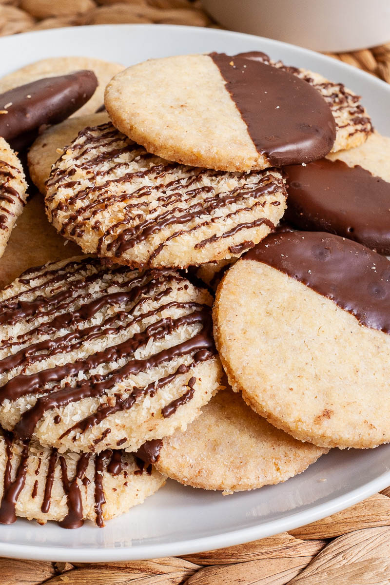 A white plate is full of thin round cookies placed on top of each other. Some cookies are partly covered in chocolate, some cookies are drizzled with chocolate so brown stripes can be seen.