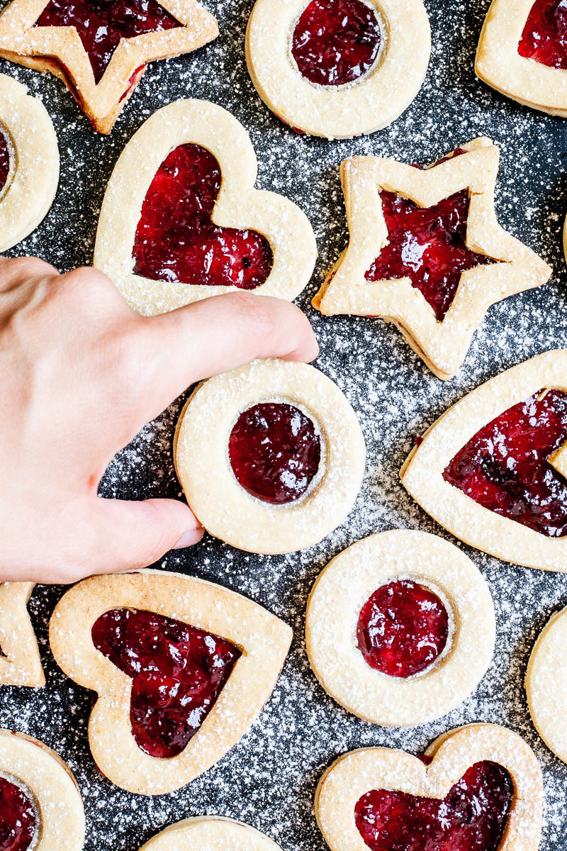 Light brown cookies in shapes of heart, star and circle filled with red jam is scattered around dusted with powdered sugar on a black surface. A hand is taking one cookie from the middle.