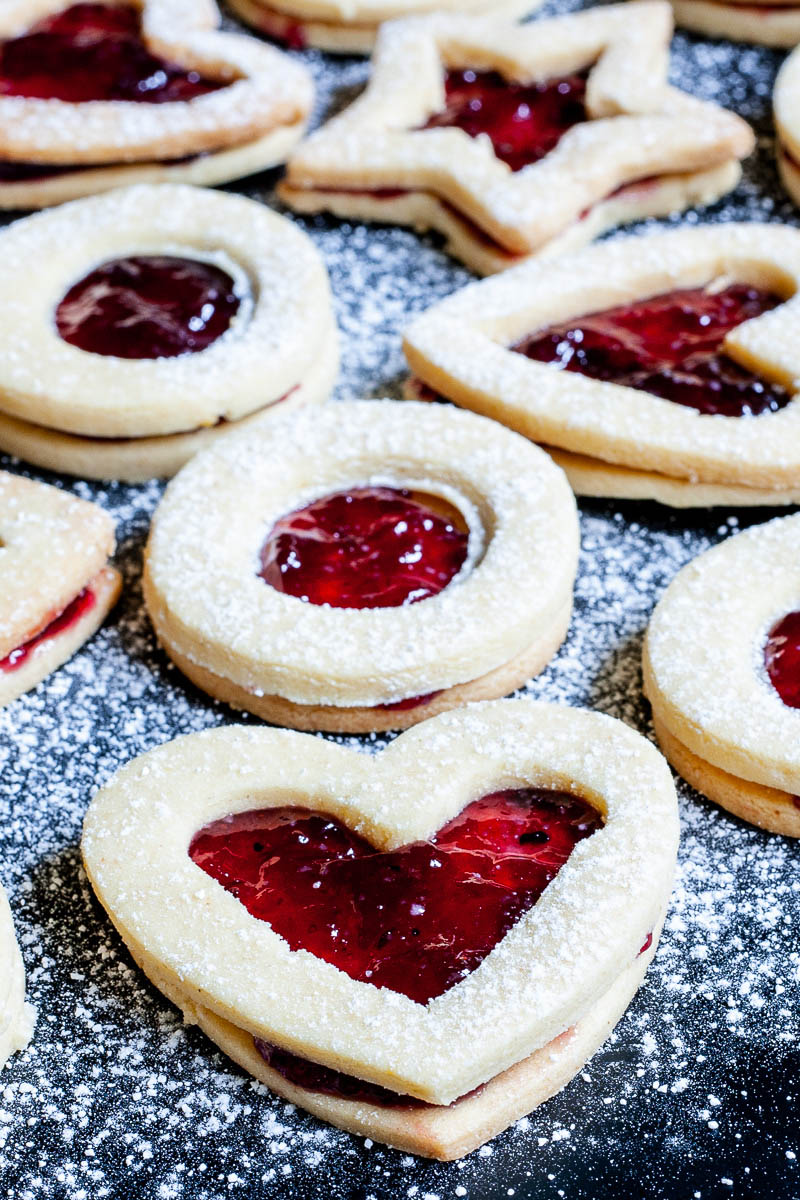 Light brown cookies in shapes of heart, star and circle filled with red jam is scattered around and dusted with powdered sugar on a black surface.