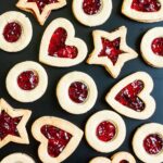 Light brown cookies in shapes of heart, star and circle filled with red jam is scattered around on a black surface.