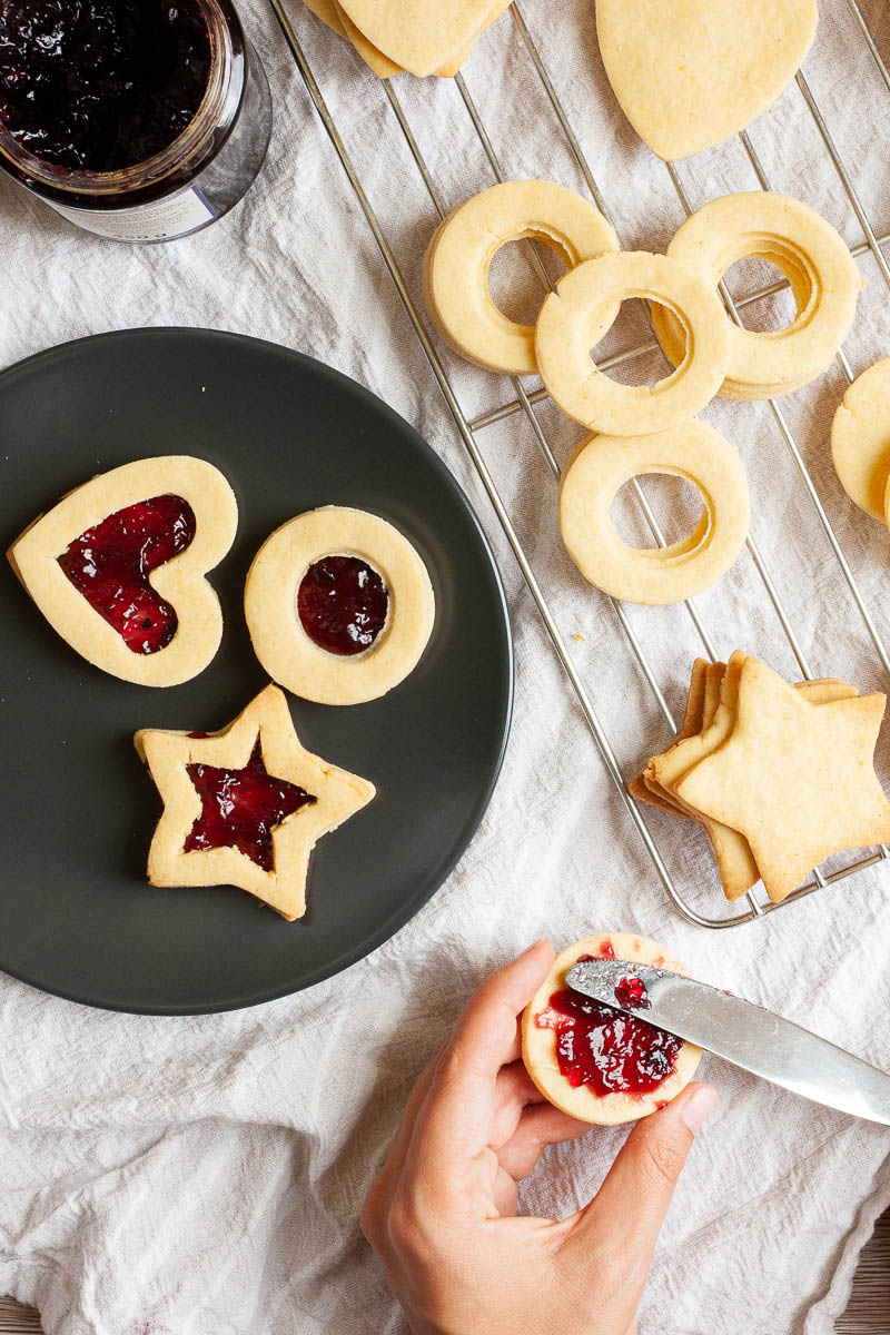 Light brown cookies in shapes of heart, star and circle are on a wire rack. A hand is holding a cookie and spreading jam on top with a knife. Jam-filled cookies are placed on a black plate.