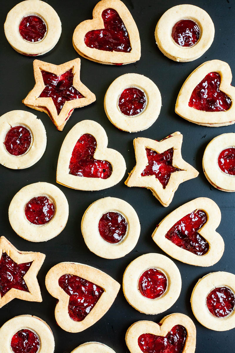 Light brown cookies in shapes of heart, star and circle filled with red jam is scattered around on a black surface.