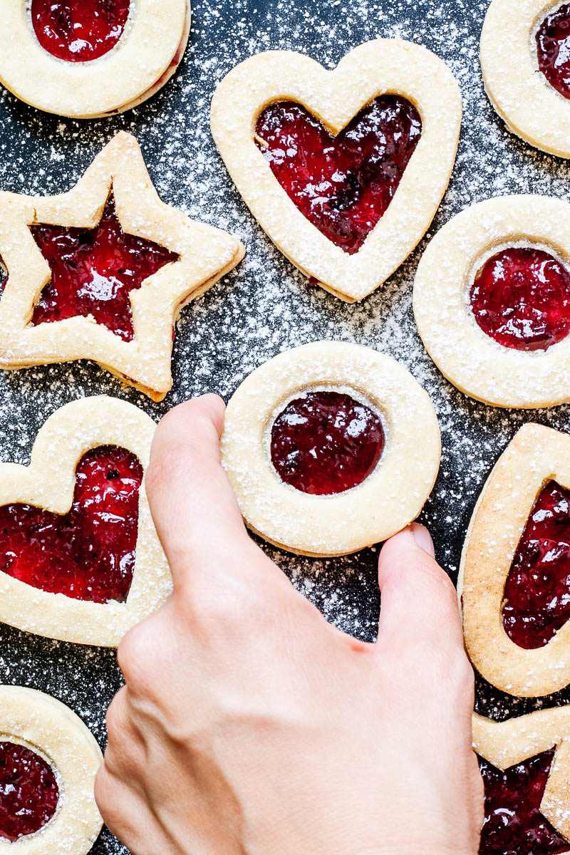 Light brown cookies in shapes of heart, star and circle filled with red jam is scattered around dusted with powdered sugar on a black surface. A hand is taking a cookie from the middle.