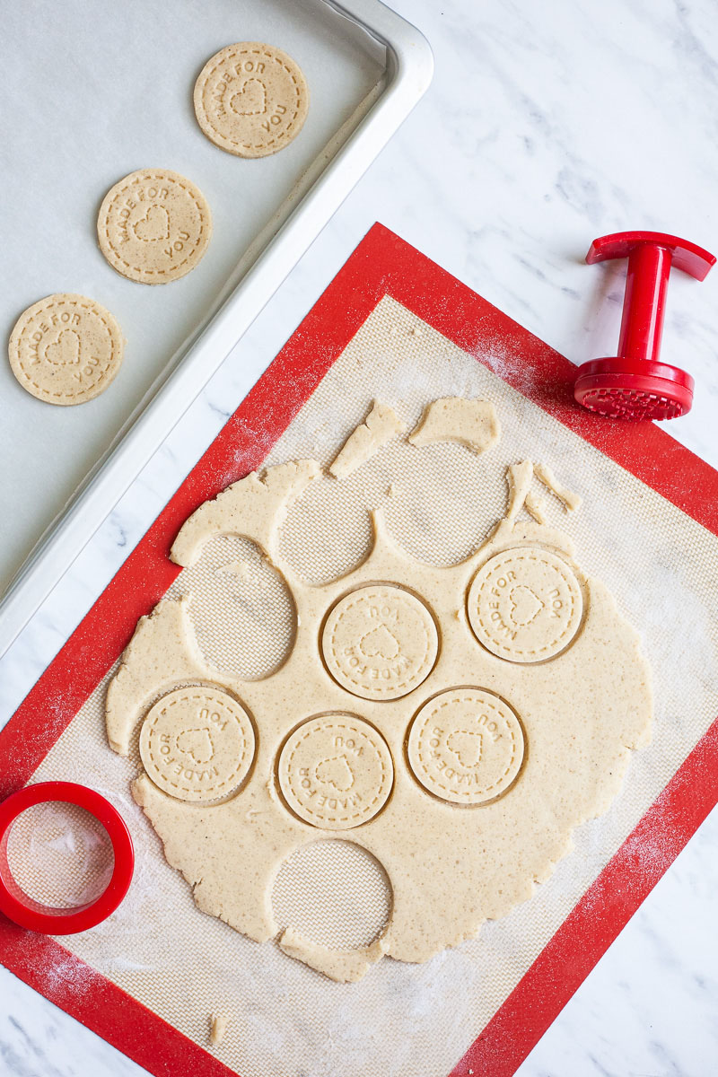 A cookie dough is rolled out on a brown-red baking mat and round cookies are cut and placed on a silver baking tray