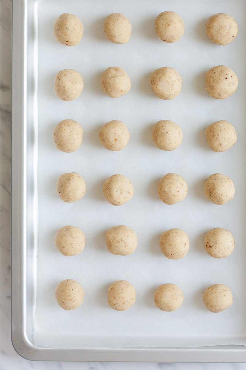 Several rows of light brown ball-shaped cookies on a silver sheet pan lined with parchment paper from above.