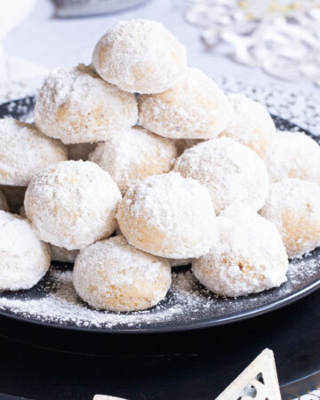 A stack of ball-shaped cookies on a black plate. It is dusted generously with powdered sugar so they are white as snow balls. White and silver christmas ornaments are placed next to them.
