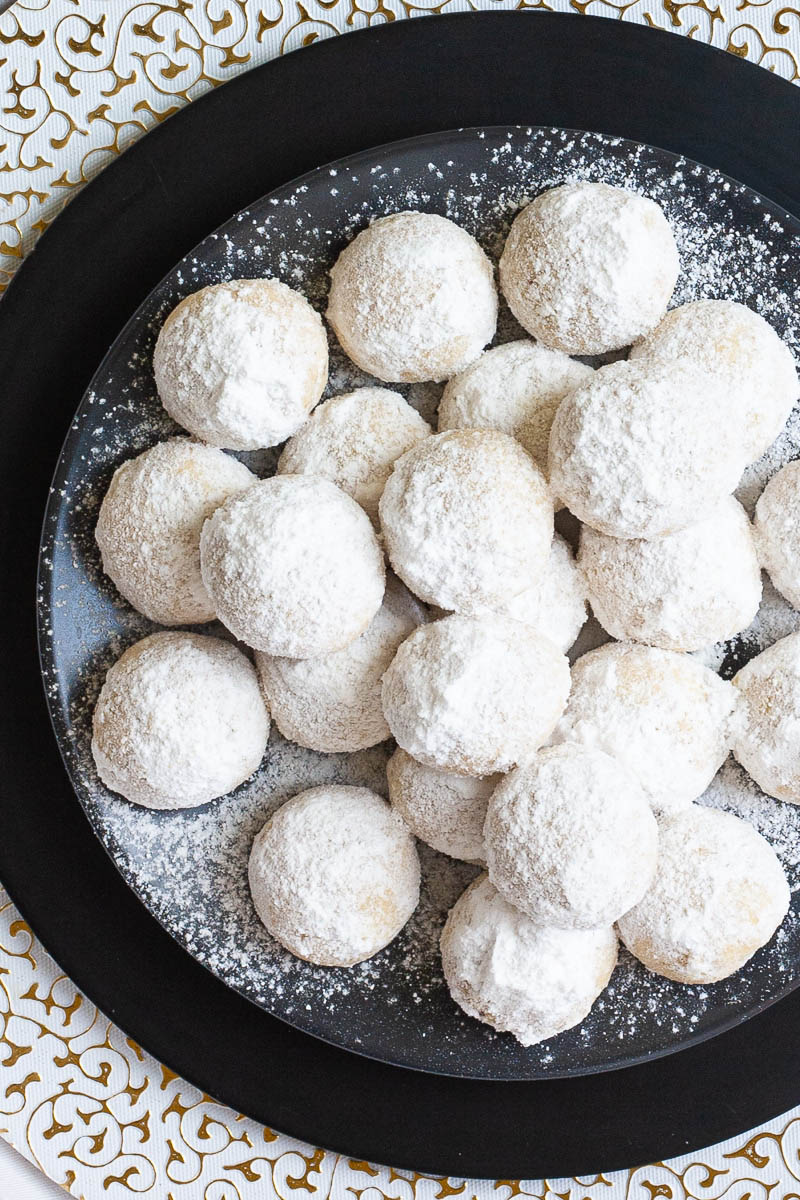 A stack of ball-shaped cookies on a black plate from above. It is dusted generously with powdered sugar so they are white as snow balls.