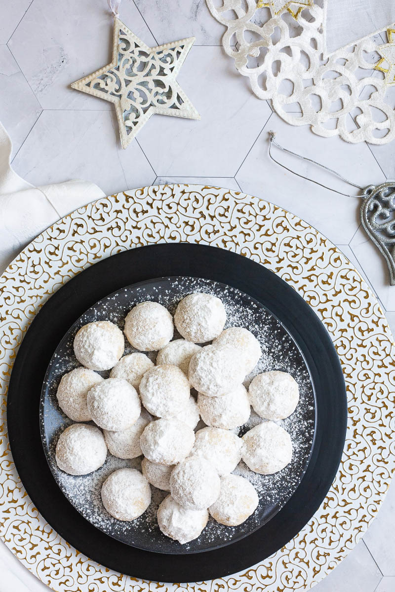 A stack of ball-shaped cookies on a black plate from above. It is dusted generously with powdered sugar so they are white as snow balls. White and silver christmas ornaments are placed next to them.