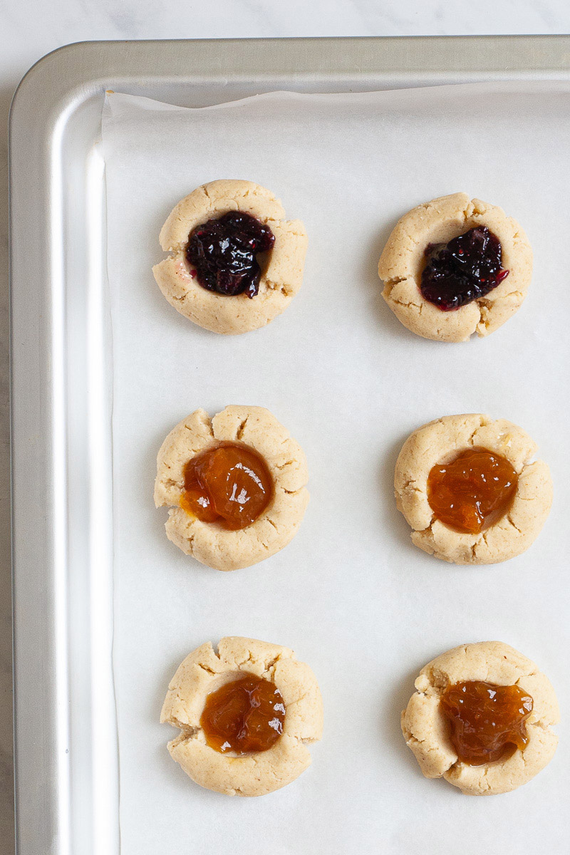 Several round cookies with orange and purple jam filling placed on a parchment paper inside a silver baking sheet ready to go to the oven.