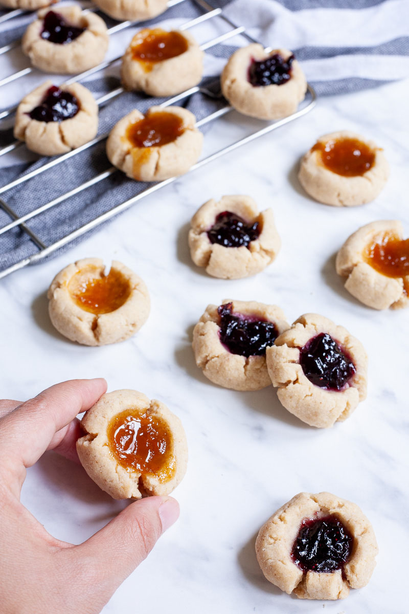 Several round cookies with orange and purple jam filling placed on a wire rack and on a marble surface. A hand is holding one cookies.