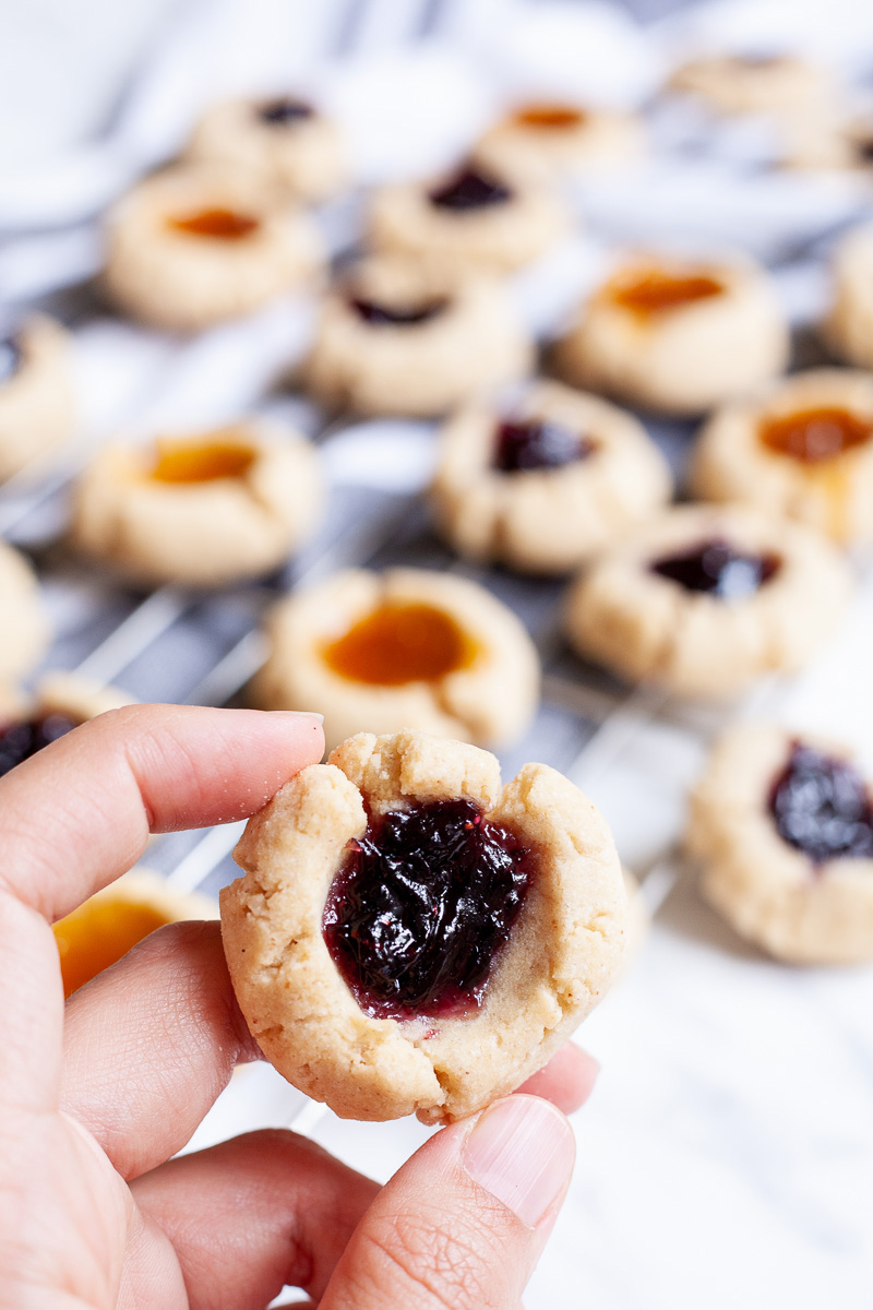 Several round cookies with orange and purple jam filling placed on a wire rack. A hand is holding one cookie up close.