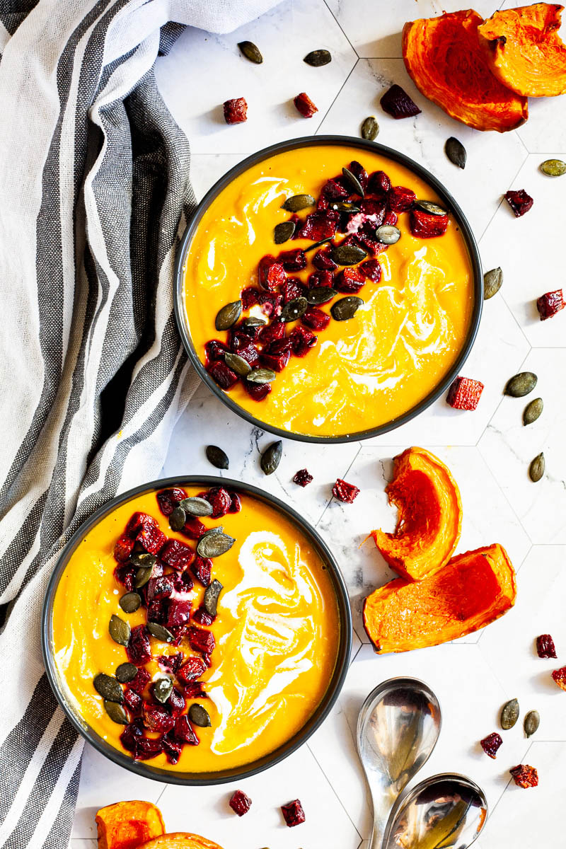Yellow cream soup in two black bowls topped with diced roasted beetroot, green pumpkin seeds and drizzled with a white sauce. Pumpkin seeds and roasted butternut squash slices are next to them.