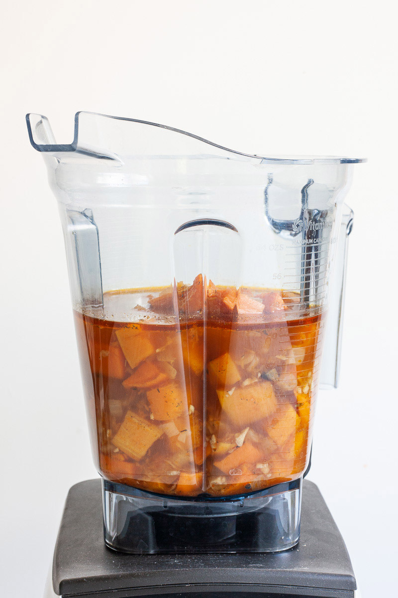 A blender from the side with red-orange soup and chopped vegetables. It is a picture before blending.