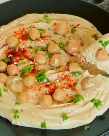 Light brown hummus dip on a black plate topped with chickpeas, chopped parsley and red paprika powder. A hand is holding a flatbread and taking a dip.