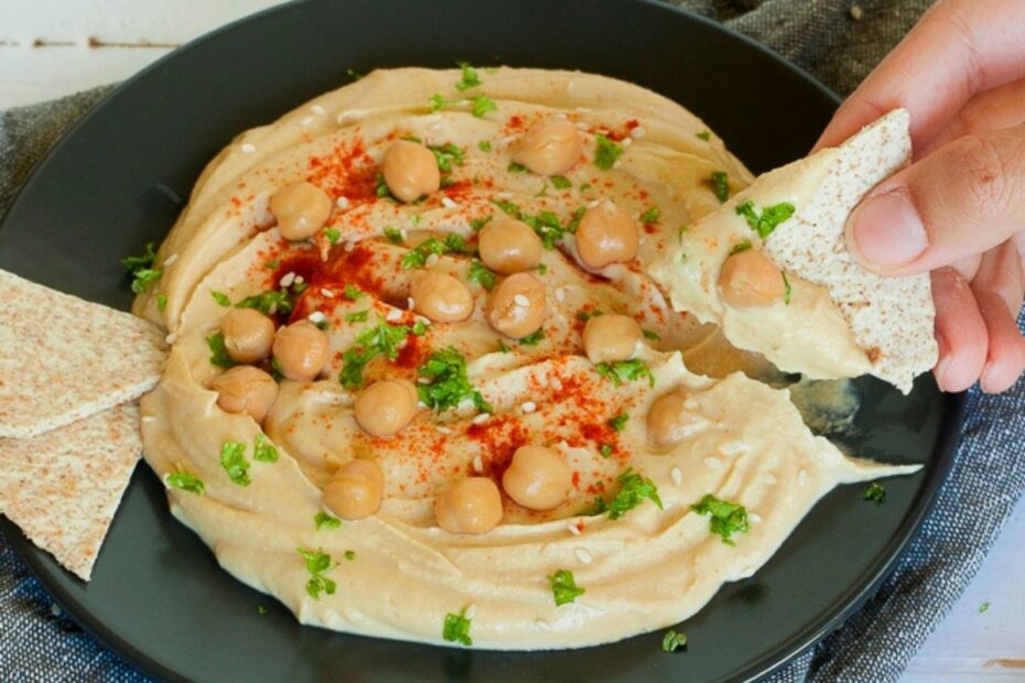 Light brown hummus dip on a black plate topped with chickpeas, chopped parsley and red paprika powder. A hand is holding a flatbread and taking a dip.