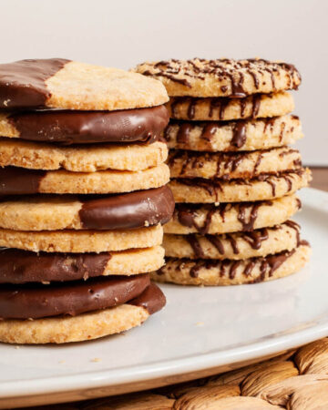 2 stack of thin round cookies on a white plate. One stack has half of the cookies covered with melted chocolate. The other stack has brown stripes of chocolate.
