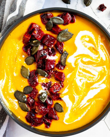 Yellow cream soup in a black bowl topped with diced roasted beetroot, green pumpkin seeds and drizzled with a white sauce.