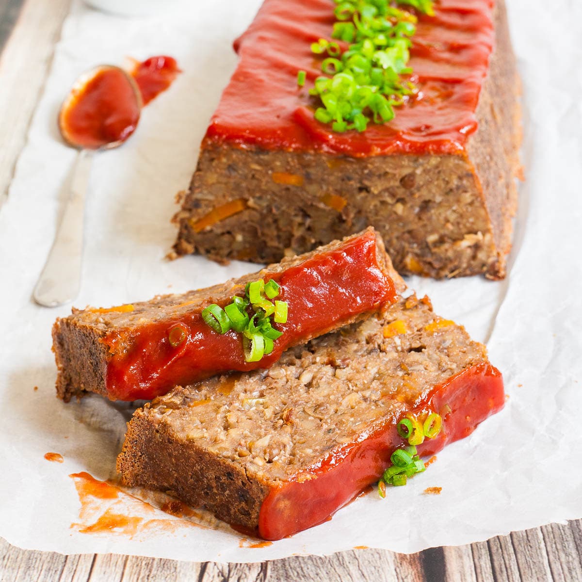 A long brown lentil loaf on parchment paper has been cut into slices. 2 slices has fallen in the front. All slices are glazed with red sauce and topped with chopped scallions.