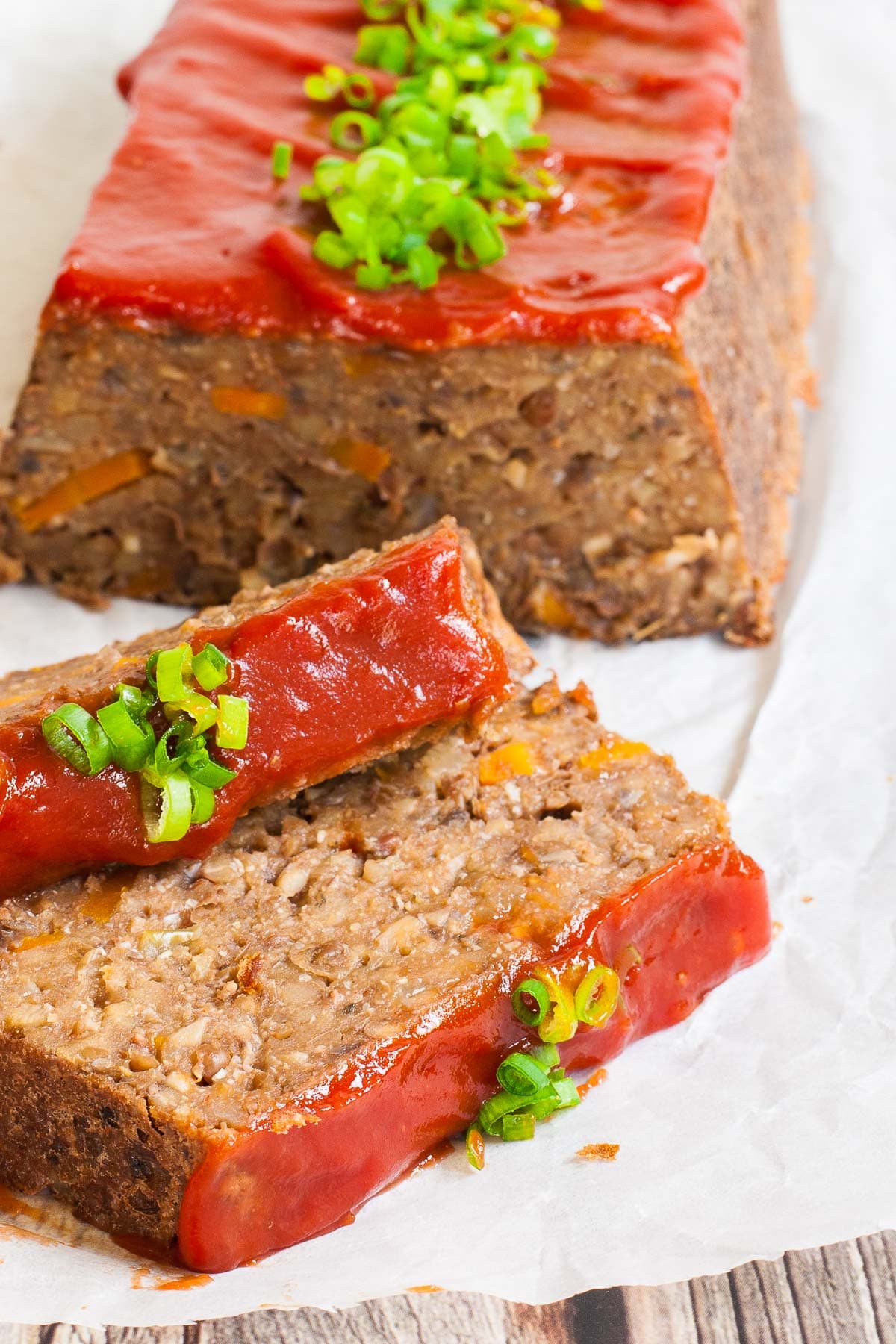 A long brown lentil loaf on parchment paper has been cut into slices. 2 slices has fallen in the front. All slices are glazed with red sauce and topped with chopped scallions.