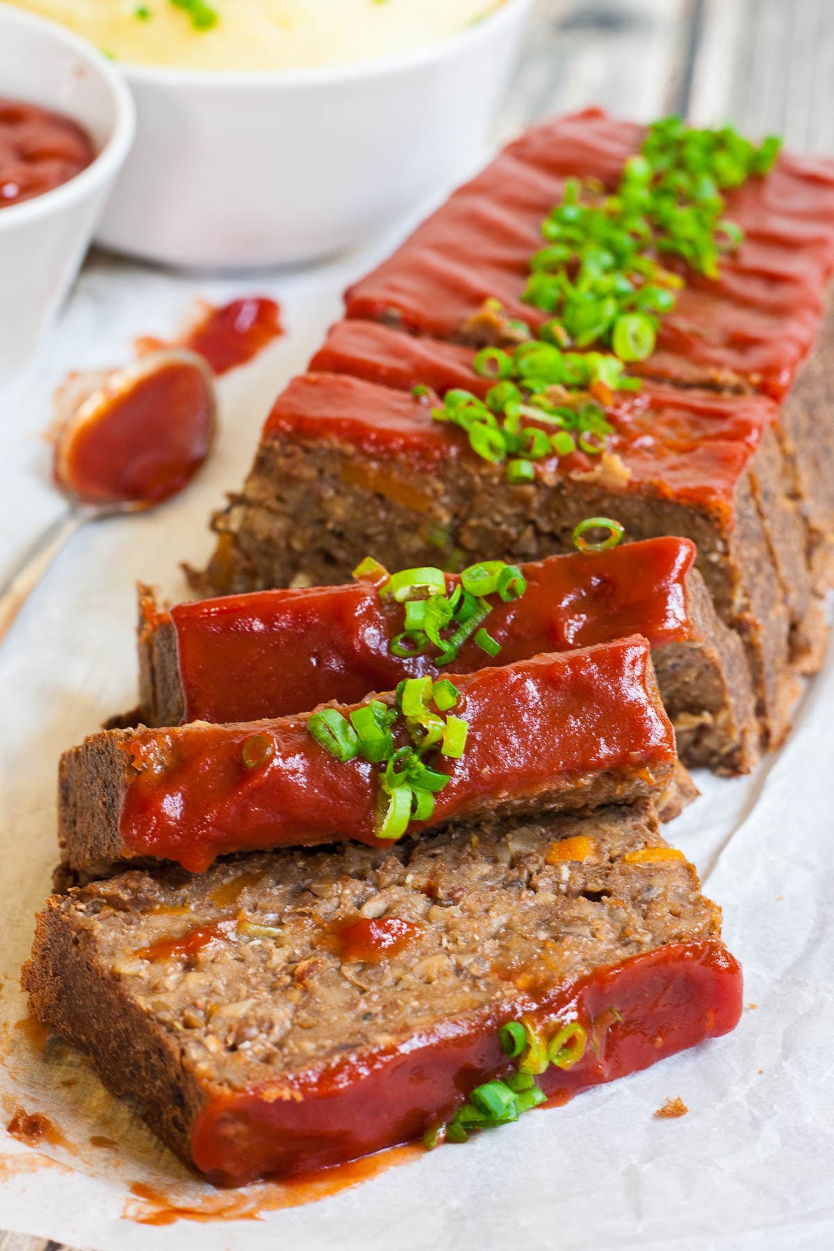 A long brown lentil loaf on parchment paper has been cut into slices. 3 slices has fallen in the front. All slices are glazed with red sauce and topped with chopped scallions. 