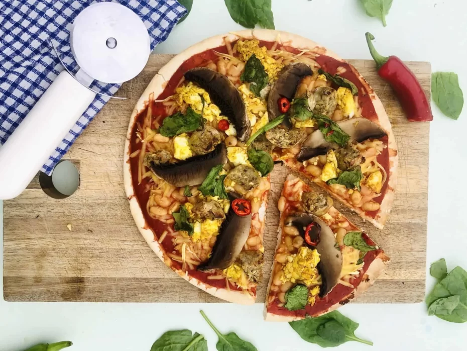 A pizza from above on a wooden board topped with mushroom slices, yellow scrambled tofu, white beans, red sauce and basil leaves