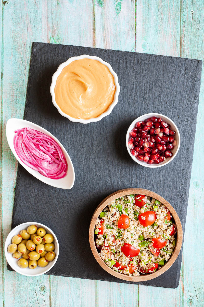 Black board with plenty of bowls with orange hummus, tabbouleh salad, pomegranate seeds, green olives and red onion