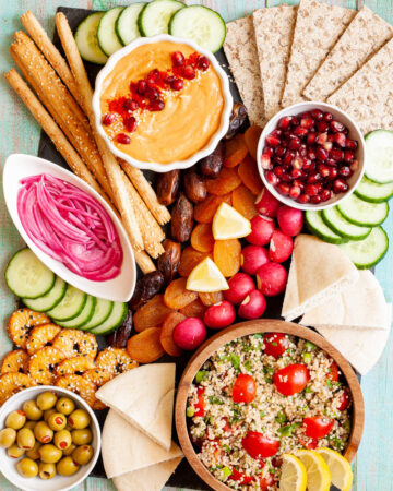 Black board with plenty of bowls, fruits and vegetables like cucumber, olives, dried apricot, pomegranate seeds, dates, orange hummus, tabbouleh salad