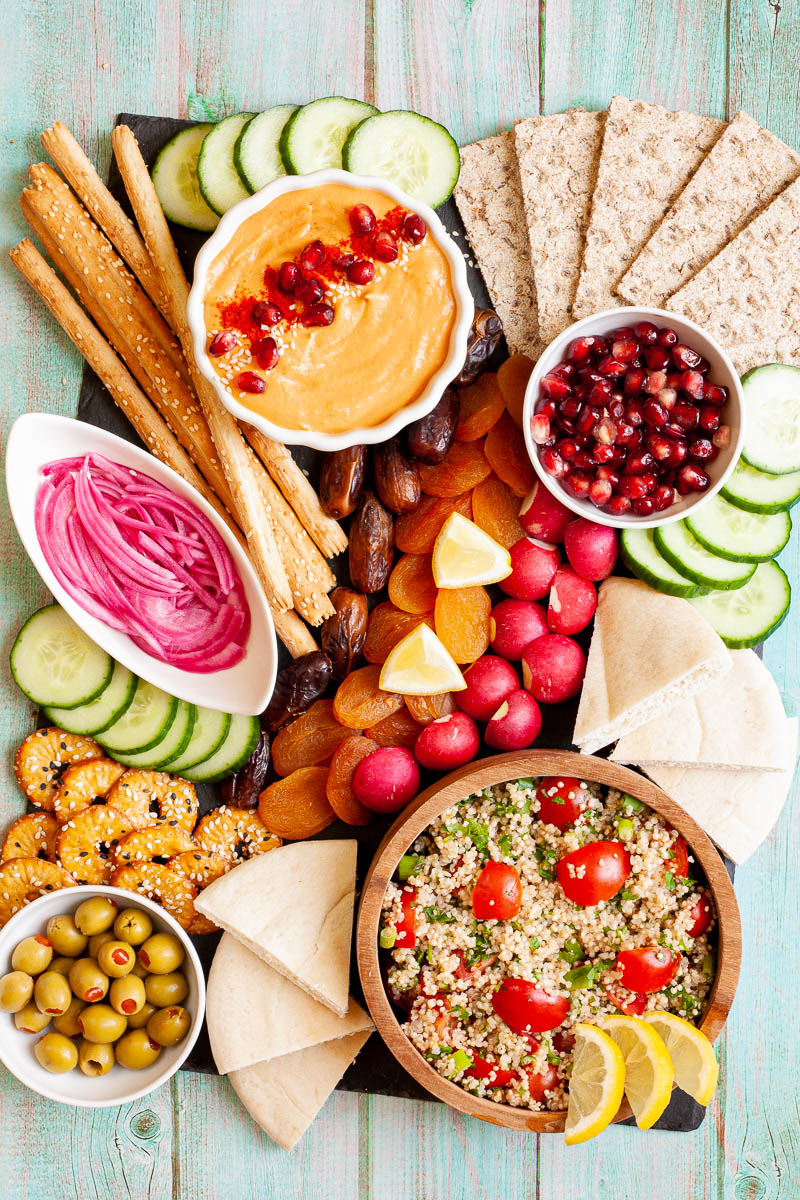 Black board with plenty of bowls, fruits and vegetables like cucumber, olives, dried apricot, pomegranate seeds, dates, orange hummus, tabbouleh salad