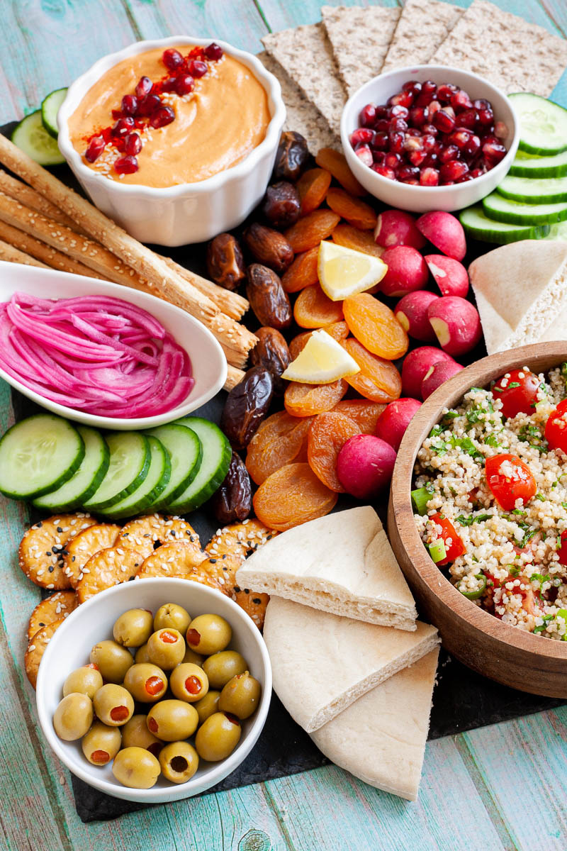 Black board with plenty of bowls, fruits and vegetables like cucumber, olives, dried apricot, pomegranate seeds, dates, orange hummus, tabbouleh salad