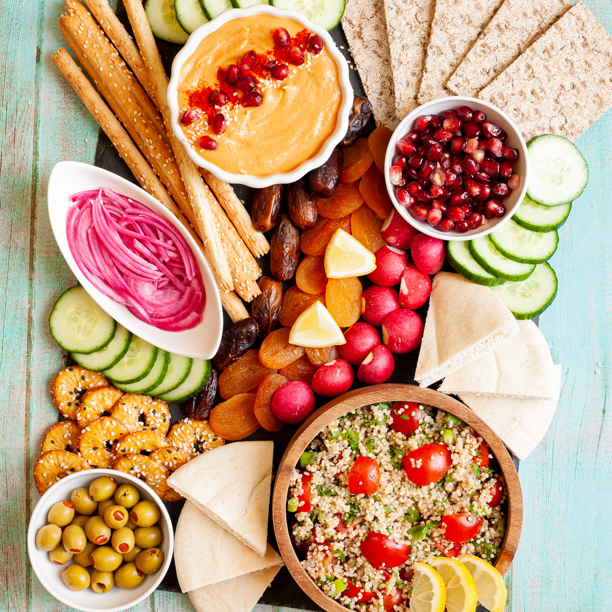 Black board with plenty of bowls, fruits and vegetables like cucumber, olives, dried apricot, pomegranate seeds, dates, orange hummus, tabbouleh salad