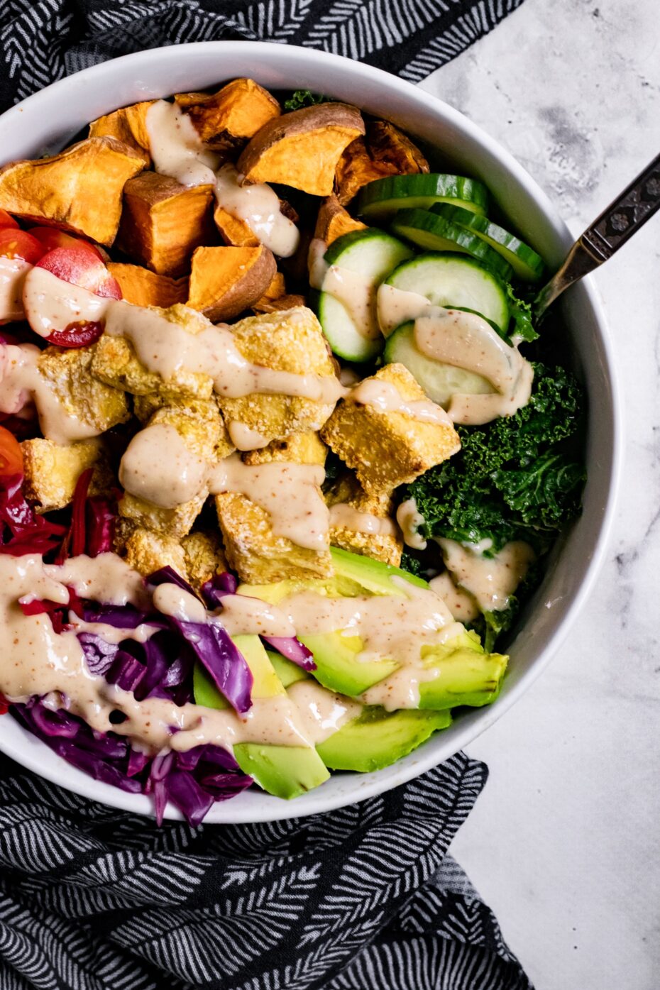 White bowl from above with avocado slices, cucumber, purple cabbage, tofu cubes and diced sweet potato  drizzled with a light brown dressing