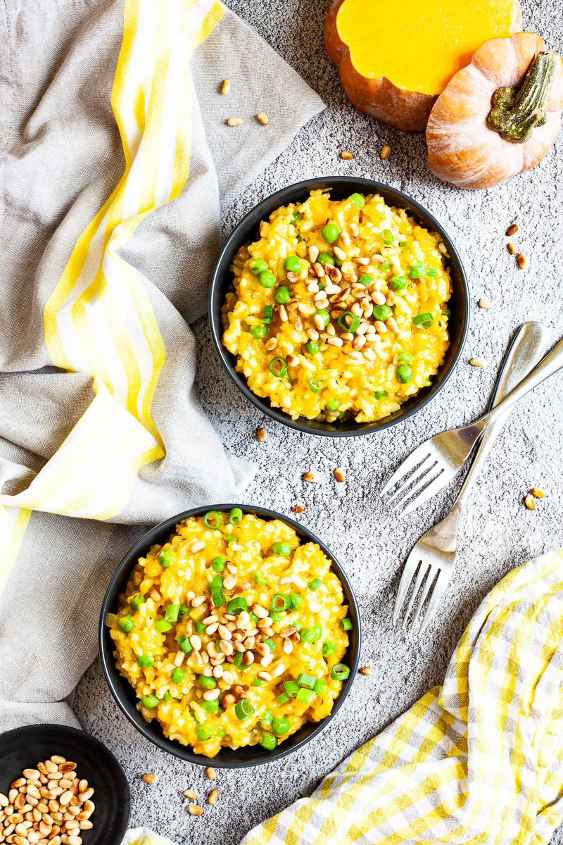 Two black bowls from above on a grey surface full of yellow risotto topped with pine nuts and chopped spring onion. Forks are placed next to them. There is some leftover raw butternut squash next to it as well as pine nuts in a small black plate.