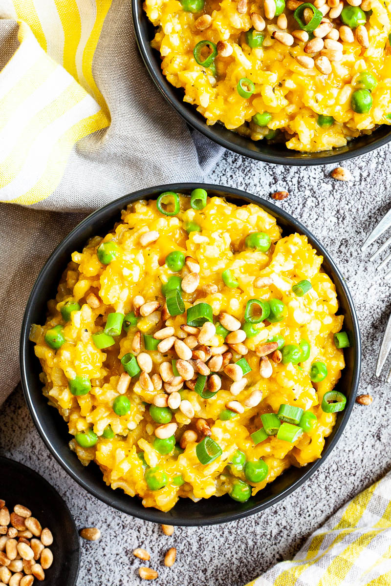 Two black bowls from above on a grey surface full of yellow risotto topped with pine nuts and chopped spring onion.