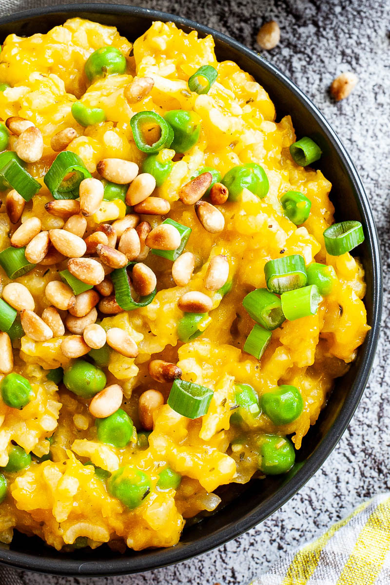 A black bowl from above on a grey surface full of yellow risotto topped with pine nuts and chopped spring onion.