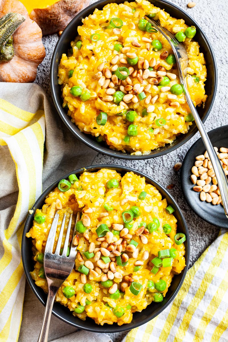 Two black bowls from above on a grey surface full of yellow risotto topped with pine nuts and chopped spring onion.  Forks are placed on them. There is some leftover raw butternut squash next to it.