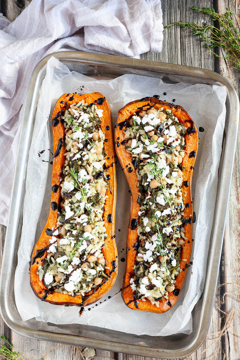 Silver baking tray from above with 2 orange butternut squash cut in half and stuffed with rice, spinach, chickpeas, olives and crumbles feta cheese drizzled with black balsamic vinegar