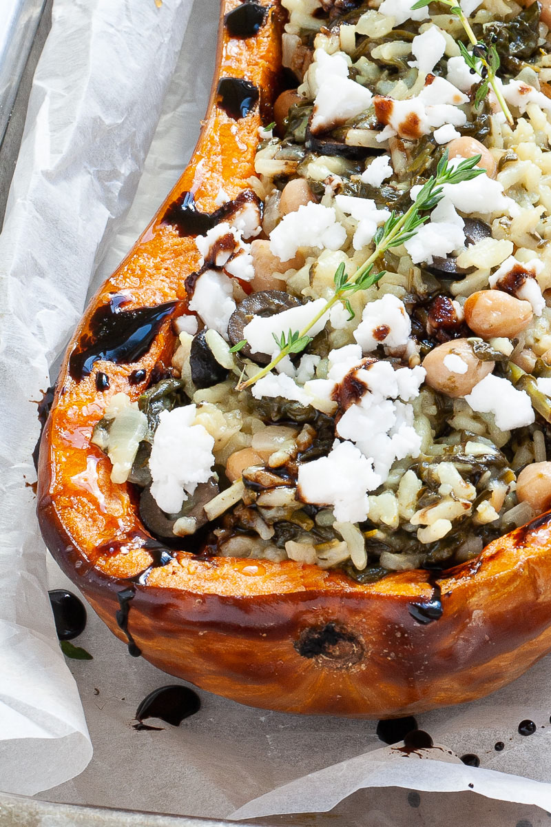 Silver baking tray with orange butternut squash cut in half and stuffed with rice, spinach, chickpeas, olives and crumbles feta cheese drizzled with black balsamic vinegar