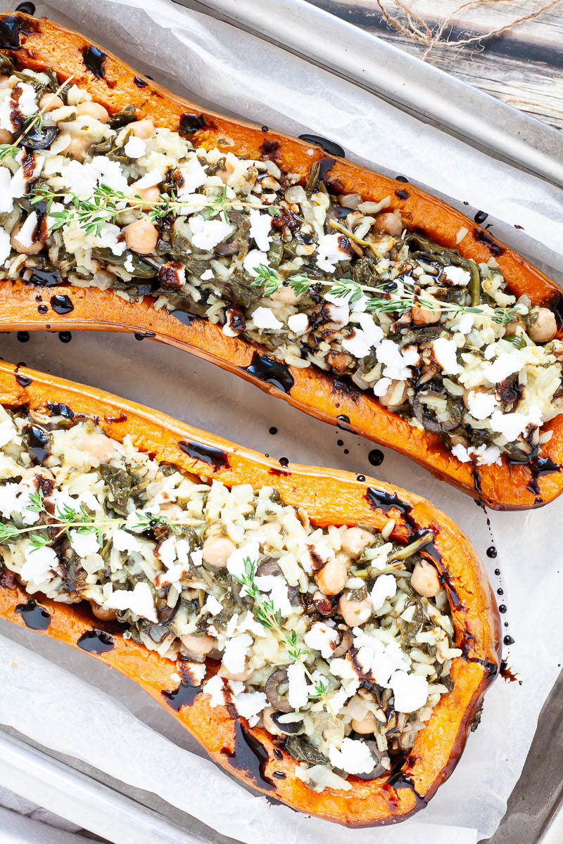 Silver baking tray with orange butternut squash cut in half and stuffed with rice, spinach, chickpeas, olives and crumbles feta cheese drizzled with black balsamic vinegar