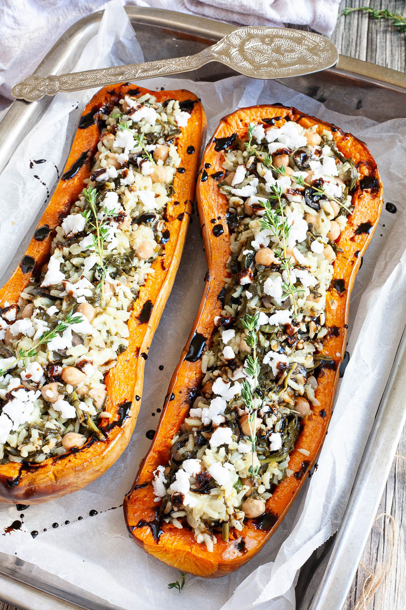 Silver baking tray from above with 2 orange butternut squash cut in half and stuffed with rice, spinach, chickpeas, olives and crumbles feta cheese drizzled with black balsamic vinegar