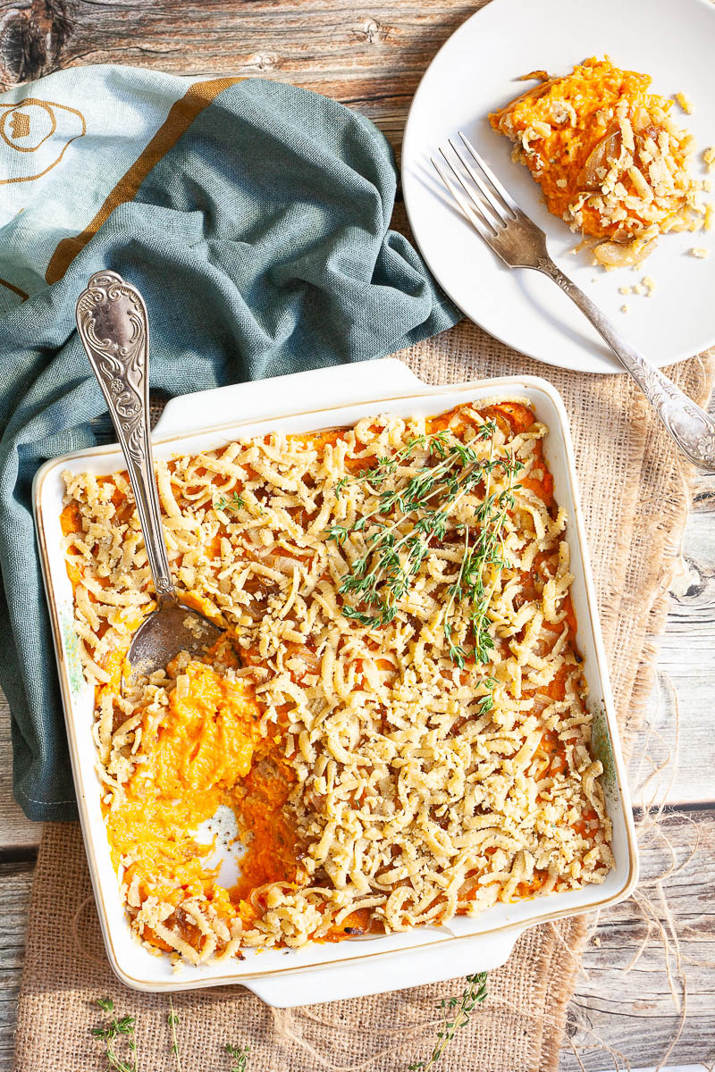 A white casserole dish from above showing orange mash topped with grated crumbles and thyme twigs. A serving spoon is dipped within. The first serving is on the background on a white plate.