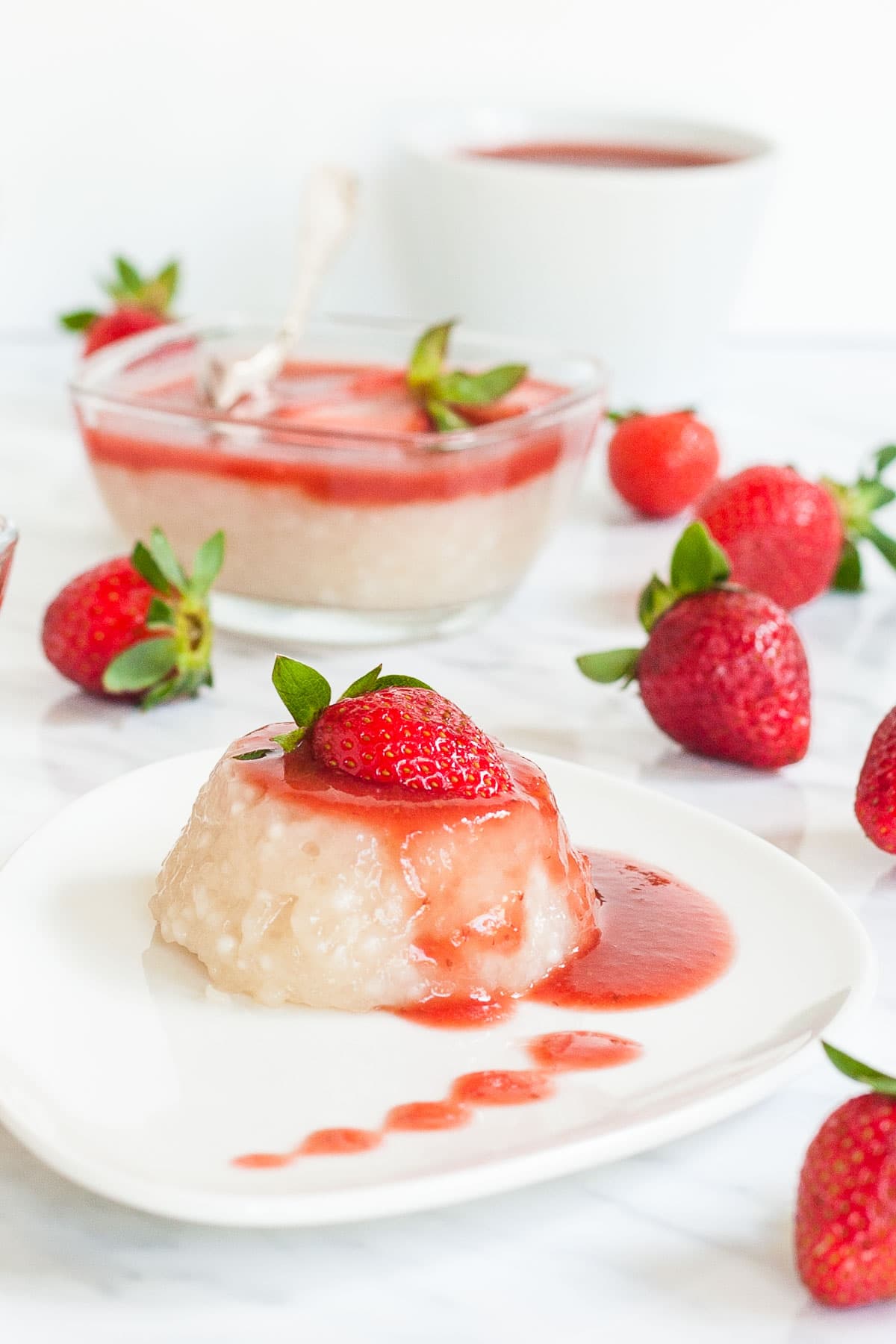 A small white plate with a heap of white pudding with pearls drizzled with strawberry sauce and topped with a half fresh strawberry. Other bowls of pudding and whole fresh strawberries are scattered around it.
