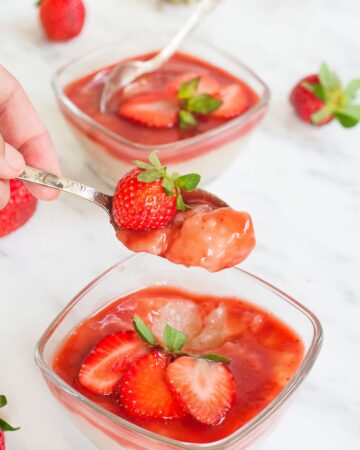 2 small glass bowls with white pudding with pearls topped with strawberry sauce and fresh strawberry slices. A hand is holding a spoon with a mouthful of the pudding.