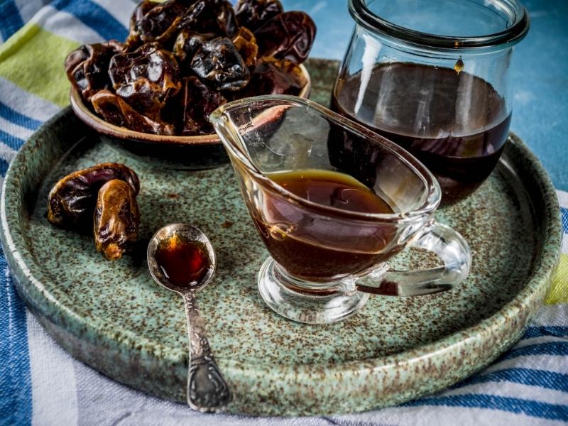 Dark brown syrup in a glass container with dates in a small wooden bowl next to it.