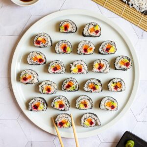 Numerous sushi rolls on a large round white plate. The rolls have black cover and white inside with purple yellow and green in the middle.