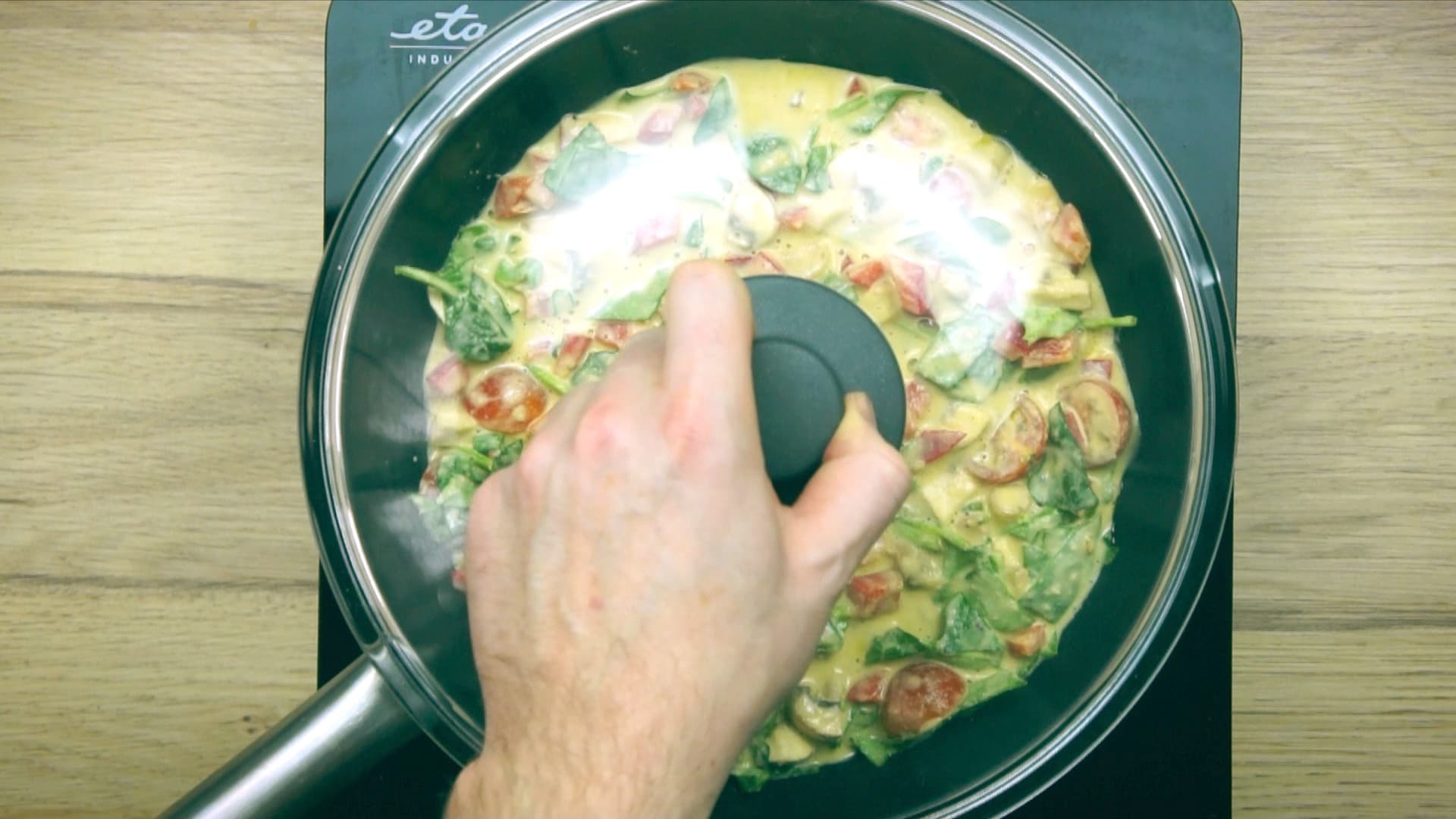 A hand is covering a yellow batter full of chopped veggies to a frying pan with a lid.